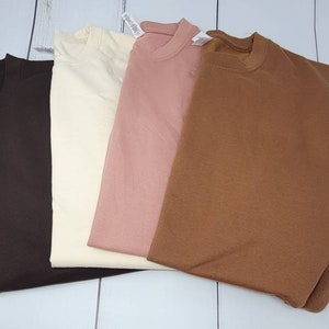 May include: Four plain, long-sleeved shirts in different colors: brown, cream, pink, and brown. The shirts are folded and laid out on a white wooden surface.