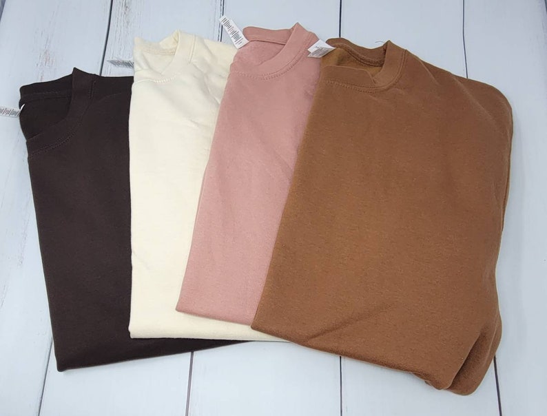 May include: Four plain, long-sleeved shirts in different colors: brown, cream, pink, and brown. The shirts are folded and laid out on a white wooden surface.