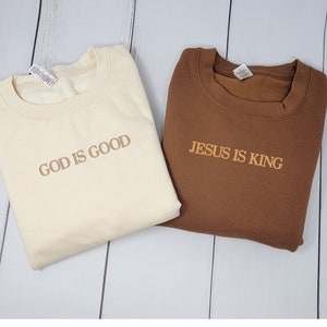 May include: Two crewneck sweatshirts with religious messages. One is beige with the text "GOD IS GOOD" in brown. The other is brown with the text "JESUS IS KING" in beige.