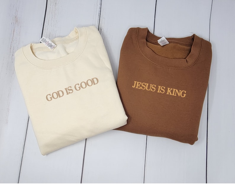 May include: Two crewneck sweatshirts with religious messages. One is beige with the text "GOD IS GOOD" in brown. The other is brown with the text "JESUS IS KING" in beige.