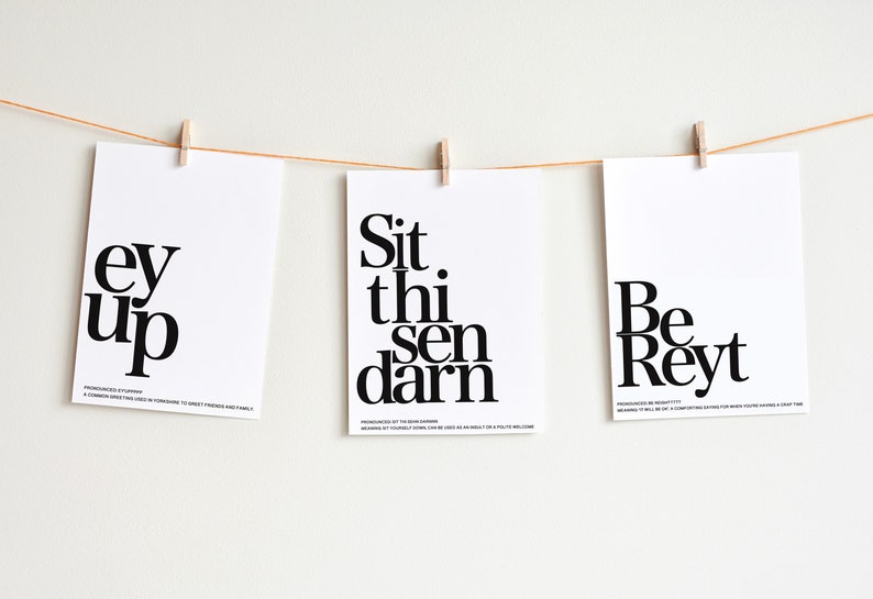 May include: Three white cards with black text hanging on a string with clothespins. The cards read "ey up", "Sit thi sen darn", and "Be Rey", each with a pronunciation guide and meaning below the text.