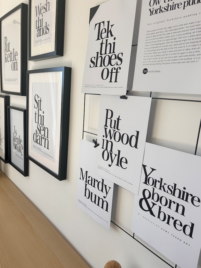 May include: A collection of black framed prints with white text on a white wall. The prints feature phrases like "Tek thi shoes off", "Put wood in oyle", "Mardy bum", "Yorkshire born & bred", "Sit thi sen darn", "Wesh thi ands", and "Is kettle brok?"