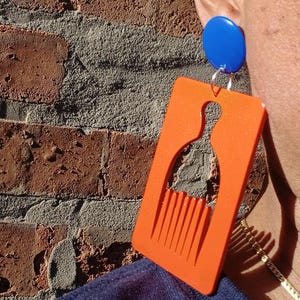 May include: An orange acrylic earring with a comb design, paired with a blue stud. The earring is rectangular with a cutout of a comb and a silhouette of a bottle. The earring is hanging from a silver hook.