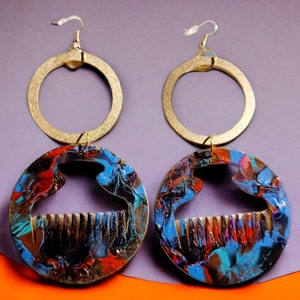 May include: A pair of gold-colored hoop earrings with a circular design. The lower portion of each earring features a round, colorful pendant with blue, red, and black paint. The pendant has a comb-like design.
