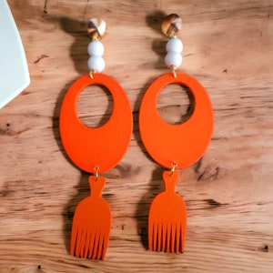 May include: A pair of orange statement earrings with a large oval shape and a smaller comb-like pendant. The earrings are made of wood and have a gold-colored metal hook.