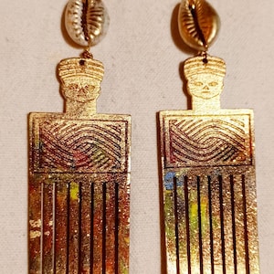 May include: A pair of gold-toned earrings shaped like hair picks, featuring a carved head at the top and a rectangular section with wavy line patterns. Each earring is accented with a white cowrie shell at the top and has a multi-colored paint finish.