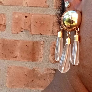 May include: Gold-toned statement earrings featuring a large, round stud and three dangling teardrop-shaped clear pendants. The earrings have a vintage aesthetic, with a polished finish. The background is a brick wall.