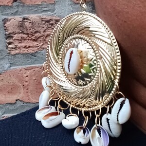 May include: Gold tone earrings with a large, round, textured disc and a dangling charm of white cowrie shells.
