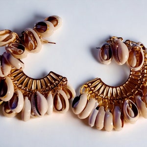 May include: A pair of gold hoop earrings with a row of small white seashells hanging from the bottom. The hoops are decorated with a gold design.