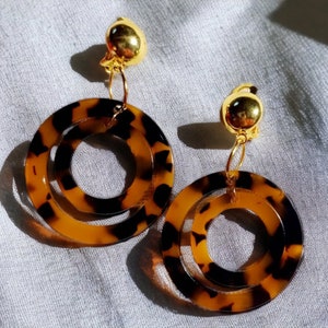 May include: A pair of gold clip-on earrings with a double-layered circle design. The circles are made of brown and black tortoise shell-like material.