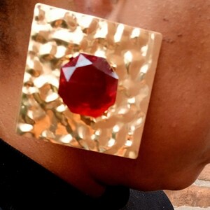 May include: Gold square earrings with a large red gemstone in the center. The earrings have a hammered texture.