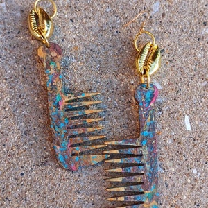 May include: A pair of handcrafted earrings featuring a comb design. The earrings have a gold-colored hook and a gold-colored shell accent. The combs are painted with a mix of blue, pink, and gold colors. The earrings are displayed on a textured surface.