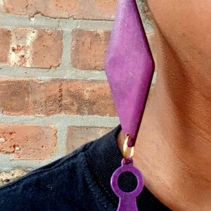 May include: A large, purple wooden earring with a geometric design. The earring features a circular stud, a diamond-shaped pendant, and a comb-shaped charm. The earring is suspended from a gold-colored hoop and is worn against a brick wall background.