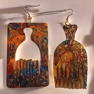 May include: A pair of colorful earrings made of wood with a marbled paint design. One earring is shaped like a comb and the other is shaped like a person with a comb for hair.