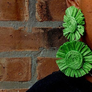 May include: Bright green, handmade earrings with a layered, floral design. The earrings are circular, with a textured, paper-like appearance. The background is a brick wall.