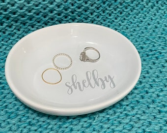 May include: White ceramic ring dish with the name "shelby" written in cursive on the front. Three rings are sitting on the dish, one gold, one silver, and one with small white beads.