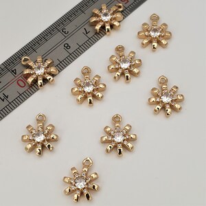May include: Ten gold-tone metal charms shaped like a flower with a clear rhinestone center. Each charm has a loop at the top for attaching to a necklace, bracelet, or other jewelry.