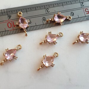 May include: Six gold-coloured jewellery charms, each featuring a teardrop-shaped, light pink gemstone and small clear accent stones. Each charm has a loop at the top. The charms are on a white surface. Ideal for jewellery making.