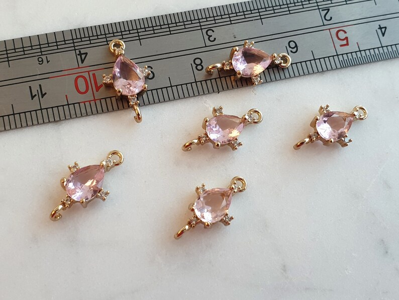 May include: Six gold-coloured jewellery charms, each featuring a teardrop-shaped, light pink gemstone and small clear accent stones. Each charm has a loop at the top. The charms are on a white surface. Ideal for jewellery making.