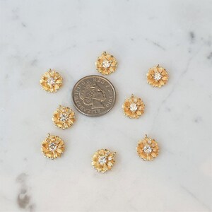 May include: Ten gold-tone flower charms with clear rhinestone centers. The charms are scattered on a white marble surface.
