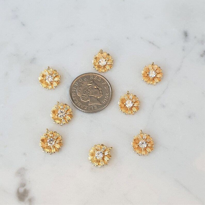 May include: Ten gold-tone flower charms with clear rhinestone centers. The charms are scattered on a white marble surface.