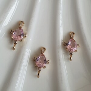 May include: Three gold-coloured teardrop-shaped charms with pink gemstones and small clear accent stones. Each charm has a small loop at the top and a hook at the bottom. The charms are displayed on a white, textured surface.
