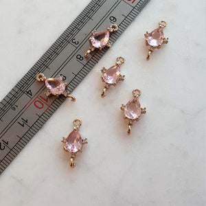 May include: A collection of gold-coloured jewellery charms. Each charm features a pear-shaped, pale pink gemstone, surrounded by small, clear accent stones. They have a loop at the top for attaching to a chain. The charms are scattered on a white surface.