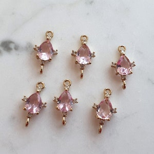 May include: Six gold-tone charms, each featuring a pink, teardrop-shaped gemstone. The charms have small, clear accent stones and a loop for attachment. They are displayed on a white marble surface.