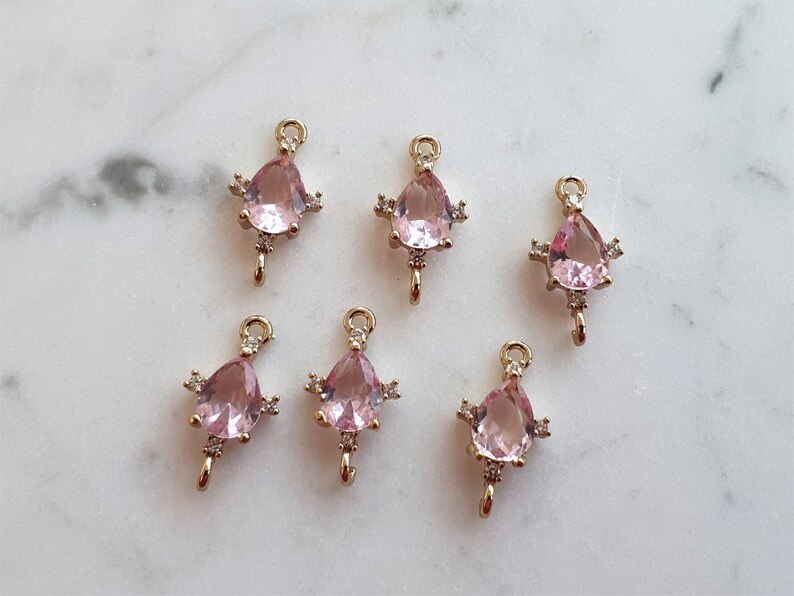May include: Six gold-tone charms, each featuring a pink, teardrop-shaped gemstone. The charms have small, clear accent stones and a loop for attachment. They are displayed on a white marble surface.