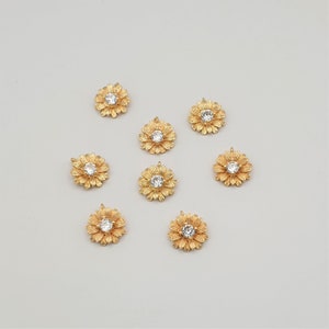 May include: Eight gold-tone flower charms with clear rhinestone centers. The charms have a loop on the back for attaching to jewelry or other crafts.