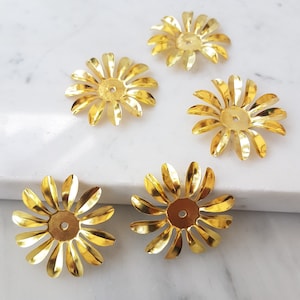 Puede incluir: Cinco adornos de metal dorado en forma de flor con un agujero en el centro. Los adornos están dispuestos en semicírculo sobre una superficie de mármol blanco.
