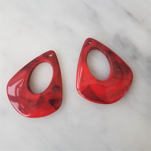 May include: Two red and black marbled teardrop-shaped earrings with a hole at the top for hanging. The earrings have a large oval cutout in the centre.