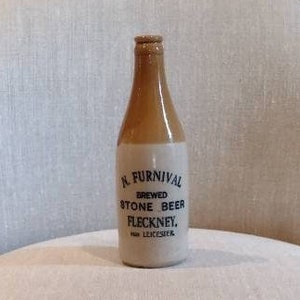 May include: A vintage stoneware beer bottle with a brown top and a cream-colored body. The bottle has the text "R. Furnival Brewed Stone Beer Fleckney, near Leicester" printed on it. The bottle is displayed on a cream-colored chair.