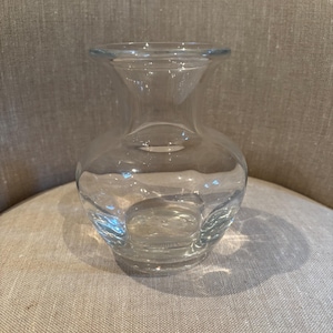 May include: A clear glass vase with a rounded body and a flared neck. The vase is sitting on a neutral-colored surface. The vase is designed to hold flowers or other decorative items.
