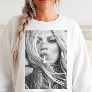 Può includere: Felpa bianca con un'immagine in bianco e nero di una donna con lunghi capelli biondi che fuma una sigaretta. L'immagine è centrata sulla parte anteriore della felpa. Il volto della donna è il punto focale.