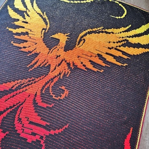 Peut inclure: Une couverture en crochet représentant un phénix qui s'élève des flammes. Le phénix est représenté dans des tons d'orange, de jaune et de rouge, tandis que les flammes sont d'un rouge vif. Le fond est noir.
