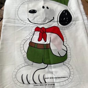 May include: A white fabric pattern for a stuffed toy of Snoopy dressed as a scout. The pattern includes a green hat, red scarf, green shorts, and a black and white outline of Snoopy's face. The text "Snoopy © 1960 United Feature Syndicate, Inc." and "Leave open for stuffing" is printed on the fabric.