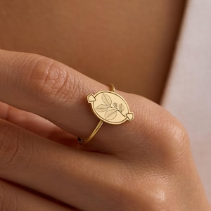 May include: A gold-colored ring featuring an oval-shaped design with a floral pattern. The ring is worn on a finger, showcasing its delicate details and elegant style. The ring's band is thin, and the oval design is centered.