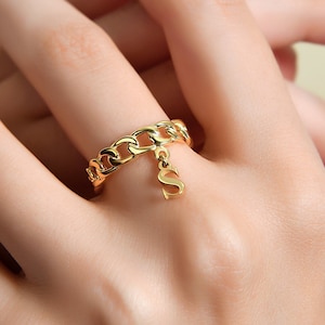 May include: Gold chain ring with a dangling letter 'S' charm. The ring is worn on a finger.
