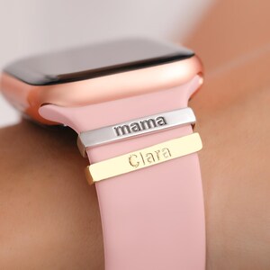 May include: A rose gold smartwatch with a pink silicone band. The band has two engraved metal bands, one silver with the word "mama" and one gold with the word "Clara".
