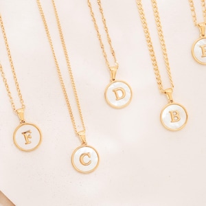 May include: Gold-toned necklaces with circular pendants, each featuring a different letter in a white background. The necklaces are arranged on a white surface with a soft, neutral-toned fabric in the background.