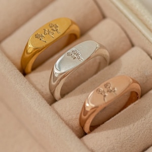 May include: Three signet rings in a ring box. The rings are gold, silver, and rose gold. Each ring has a floral design engraved on the top.