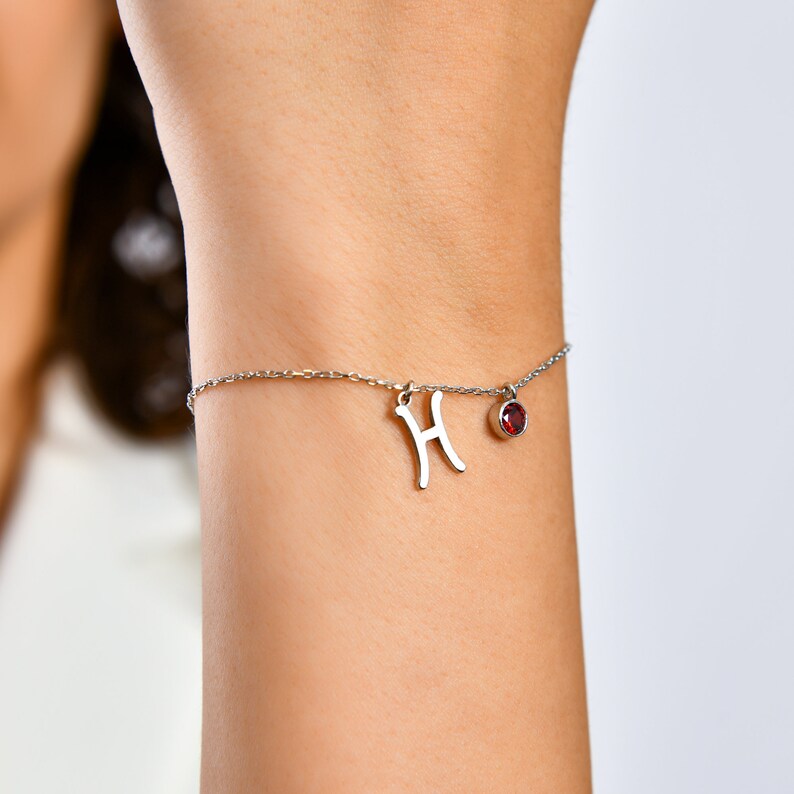 May include: A silver chain bracelet with a silver charm in the shape of the letter "H" and a small red gemstone.