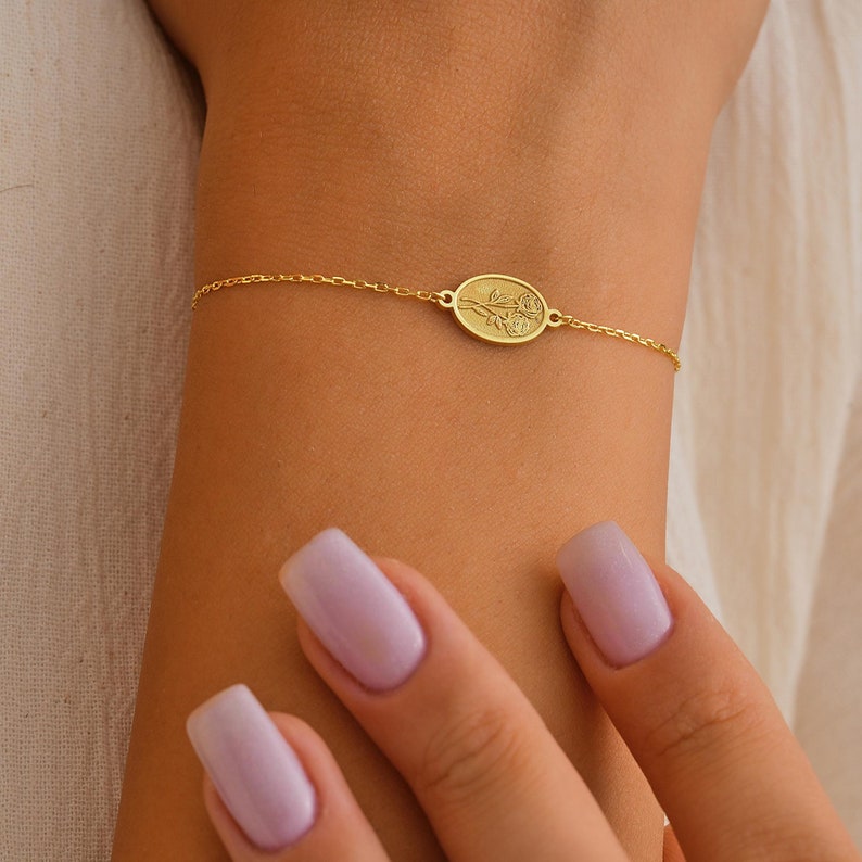 May include: A delicate gold chain bracelet with a small oval charm featuring an engraved floral design.