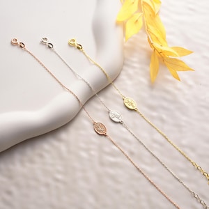 May include: Three delicate silver chain bracelets with oval charms featuring a floral design. The bracelets are displayed on a white surface with a yellow leaf branch in the background.