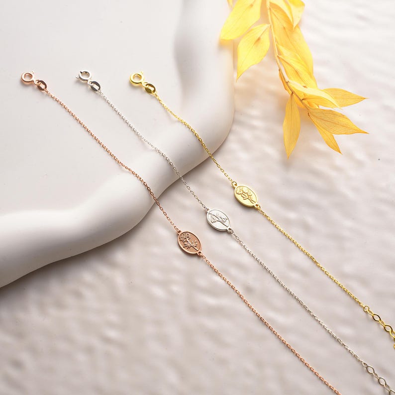 May include: Three delicate silver chain bracelets with oval charms featuring a floral design. The bracelets are displayed on a white surface with a yellow leaf branch in the background.