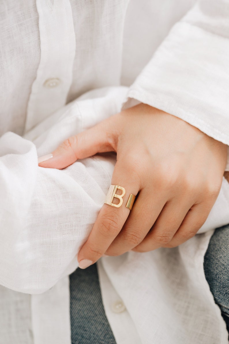 May include: A gold initial ring with the letter "B" on the band. The ring is worn on the index finger of a hand.