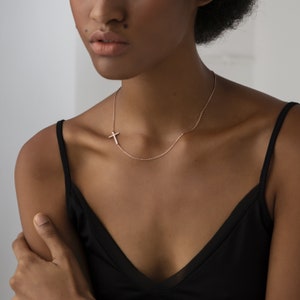 May include: A delicate gold necklace with a small cross pendant. The necklace is worn by a person with dark skin and short curly hair. The person is wearing a black tank top.