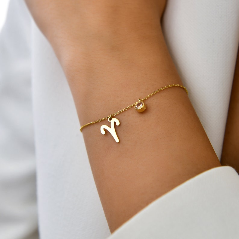 May include: A gold chain bracelet with a small clear crystal charm and a gold Aries zodiac symbol charm.
