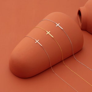 May include: Four delicate necklaces with a simple cross pendant. The necklaces are silver and gold and are displayed on a terracotta background.